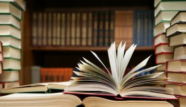 A few books lay on a table in front of a full book shelf. On the table, one book is laying open with the pages standing open between the covers.