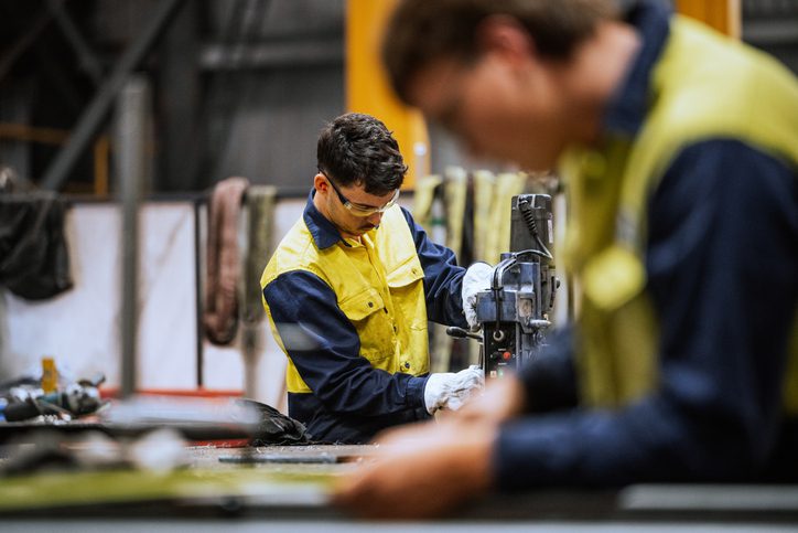 Two individuals in yellow and blue work uniforms in an industrial workshop; one operates a machine while the other is blurred in the foreground, surrounded by tools and equipment.