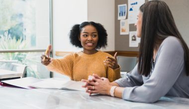 Two women are sitting at a table. While looking at each other, one has her arms open as if too demonstrate distance from one point to another.