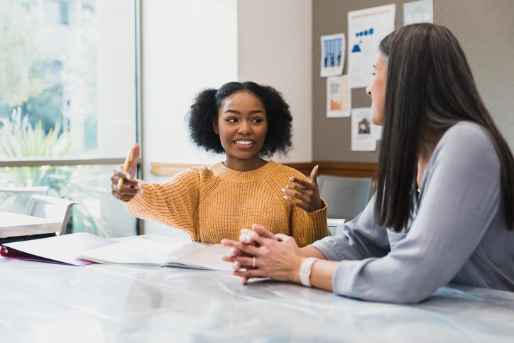 Two women are sitting at a table. While looking at each other, one has her arms open as if too demonstrate distance from one point to another. 