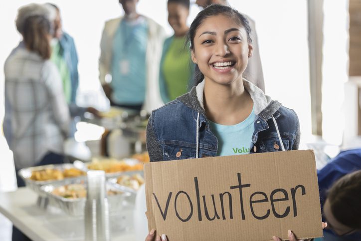 Woman holding a cardboard sign that says 'volunteer'. In the background, several people are gathered near tables with food, suggesting a community service or volunteer event.