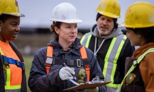 Four construction workers in safety gear gathered around a clipboard, discussing plans at a worksite. Faces are blurred for privacy.