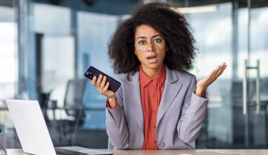 A professional woman with an afro hairstyle dressed in business attire, is holding a cellphone while in front of a laptop.