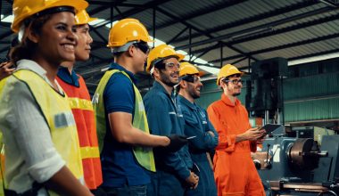 A group of workers in safety gear including helmets and reflective vests standing inside an industrial manufacturing plant with machinery in the background. One individual in an orange jumpsuit appears to be holding a tablet.