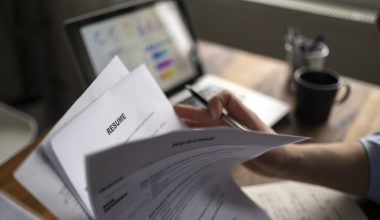 Person holding a resume and a pen in front of a laptop with a coffee mug on the desk