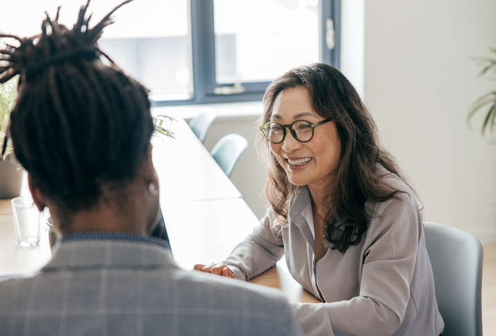 Two individuals sitting at a table in a bright office space, engaging in a conversation with one person's back towards the camera, revealing a male with dreadlocks in a patterned shirt, and the other person wearing a white blazer with dark hair, gestures suggesting a discussion or meeting is taking place.
