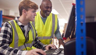 Two men in safety vests, one younger and one older, smiling and looking at a computer screen in a warehouse setting.