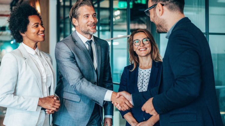 Four business professionals, three men and one woman, are engaged in a friendly handshake and conversation in a professional office setting. Two people are shaking hands while the others smile.