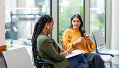 Two women sitting in chairs having a conversation while one takes notes on a clipboard.