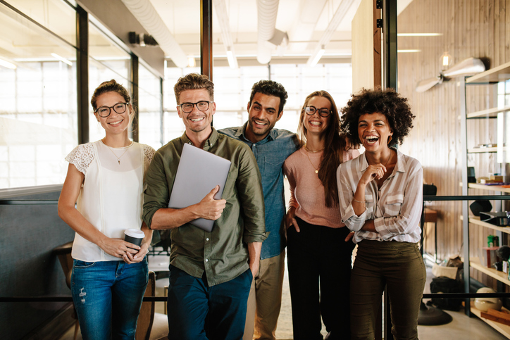 Group of five diverse coworkers smiling and posing together in a modern office environment, with two men and three women, one holding a laptop and another holding coffee cups.