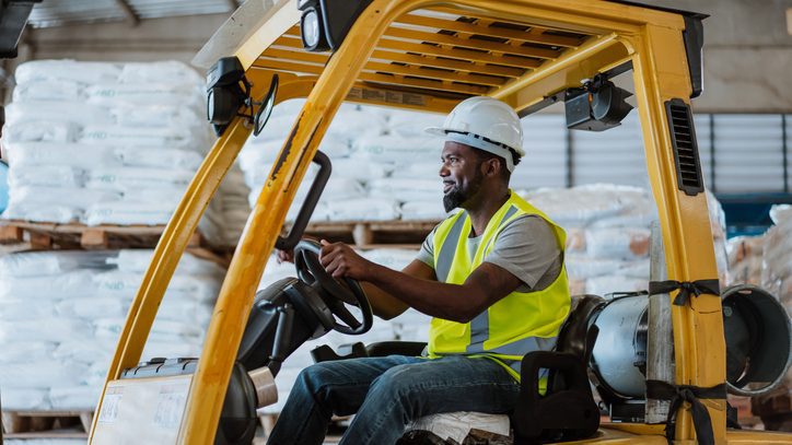 Person operating a yellow forklift in a warehouse, surrounded by stacked pallets wrapped in white plastic, wearing a hard hat and reflective vest.