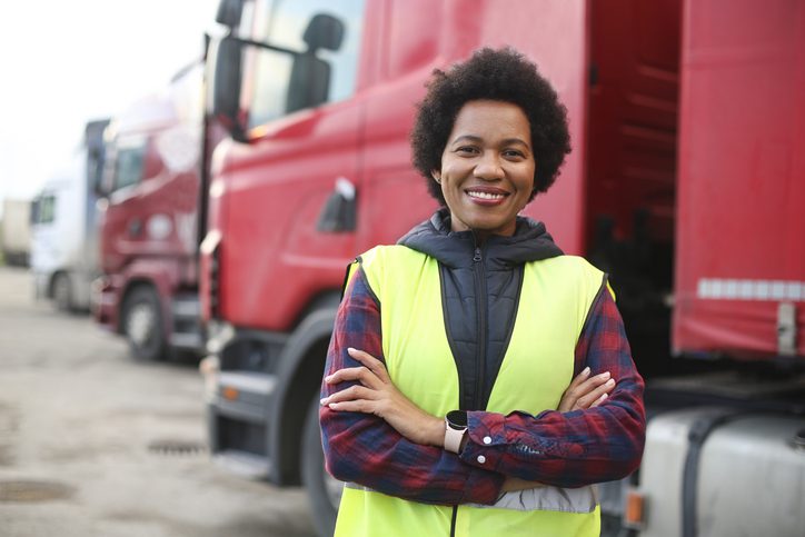 A black woman is wearing a safety vest and standing in front of a row of large parked trucks.