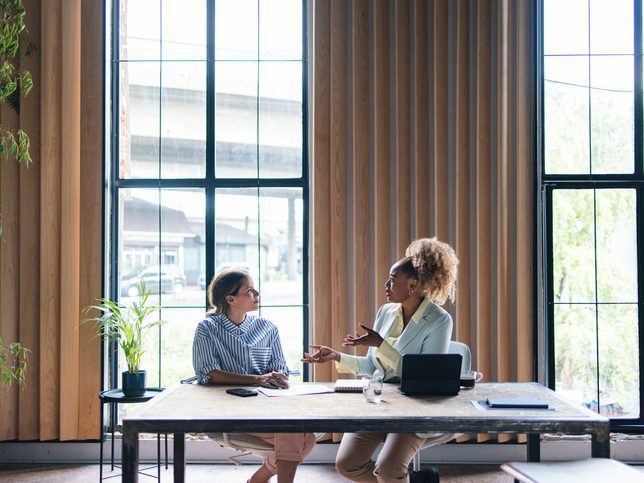 Two women having a discussion at a table in front of large windows in an office space.