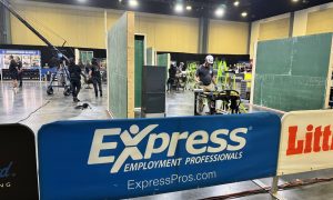 Indoor space with people working on construction tasks. Green partition walls, ladders, and tools are visible. A blue banner in the foreground reads 'Express Employment Professionals' and 'ExpressPros.com'.