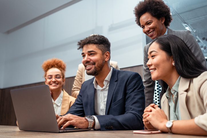 Group of professionally dressed people smiling and looking at a laptop in an office setting.