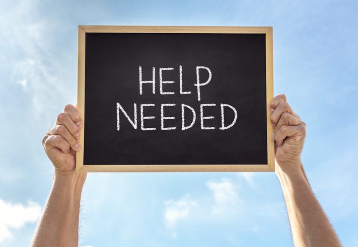 Two hands holding a small blackboard with the words 'HELP NEEDED' written in white chalk, against a clear blue sky with scattered clouds.