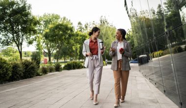 Two women walking and talking on a paved path surrounded by trees and a glass building.