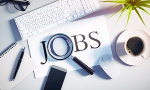 Workspace with the word "JOBS" under a magnifying glass, surrounded by a keyboard, mouse, coffee, plant, smartphone, and notepad.