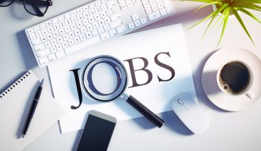 Workspace with the word "JOBS" under a magnifying glass, surrounded by a keyboard, mouse, coffee, plant, smartphone, and notepad.