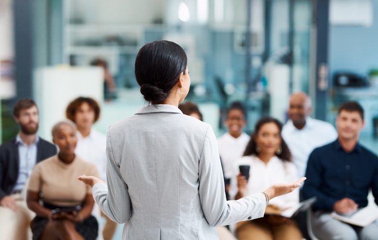 A woman in a gray blazer speaks to a seated audience in a modern office setting.