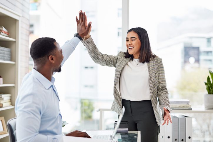 Two people in an office exchanging a high-five, with a desk and laptop in the foreground.