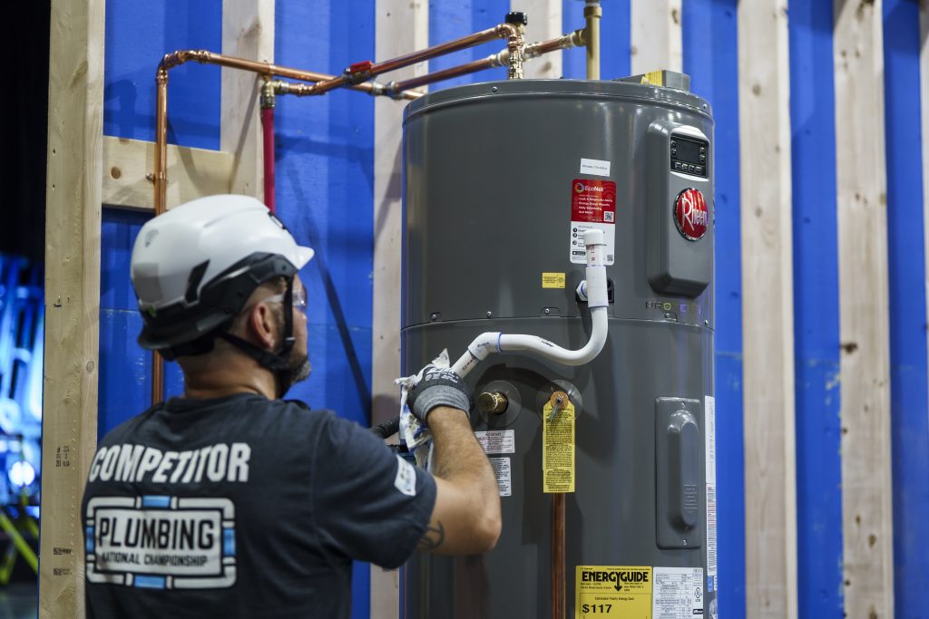 Person in safety gear working on a gray water heater against a blue and wood backdrop.