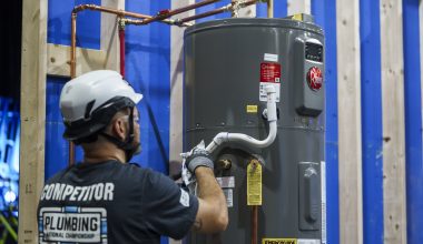 Person in safety gear working on a gray water heater against a blue and wood backdrop.