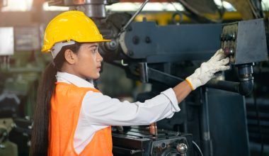 Woman in a hard hat, safety vest, and gloves operating machinery in a factory.