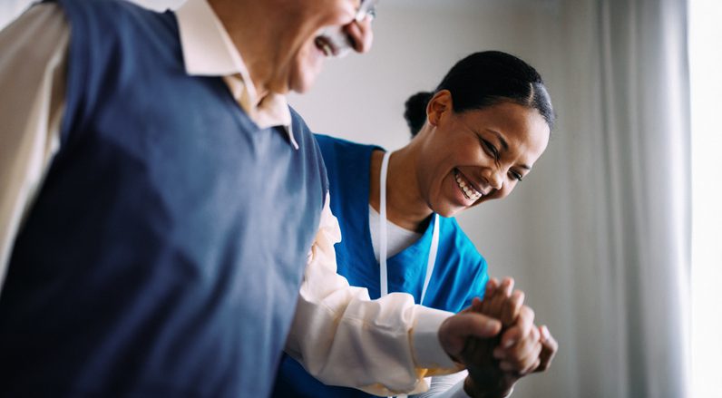 An elderly man wearing glasses and a dark vest over a white shirt is holding hands with a healthcare worker dressed in blue scrubs. The healthcare worker's face is blurred, and they appear to be assisting the elderly person. They are indoors, near a window with light curtains.