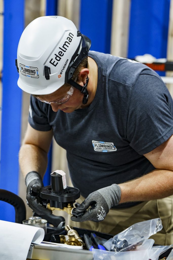 Person wearing a white hard hat labeled 'Edelman' and gloves, working with plumbing tools and equipment.