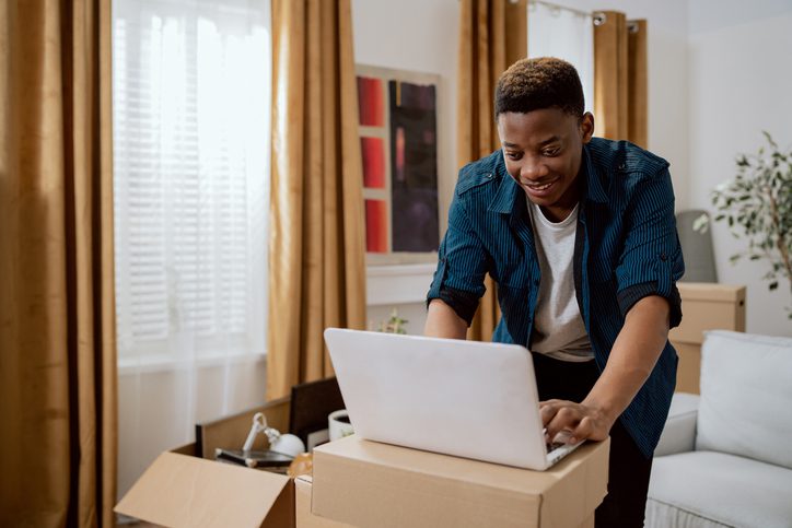 Black person working on a laptop placed on cardboard boxes in a room with golden curtains, surrounded by furniture, plants, and wall art.