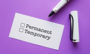 A white card with two checkboxes labeled "Permanent" and "Temporary" is placed on a purple surface. Next to the card, there is a white pen with a black tip and a white cap attached to it by a black string.