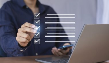 Person in a dark blue shirt using a laptop and holding a smartphone, interacting with a virtual checklist featuring five items marked with checkmarks.