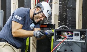 Electrician wearing helmet and gloves working on wiring inside an electrical panel, with visible colored wires.