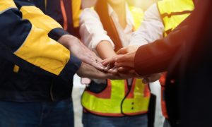 Group of workers in high-visibility safety vests and uniforms standing in a circle with hands stacked together, symbolizing teamwork, unity, and collaboration in an industrial or construction setting. The image reinforces themes of workplace solidarity, team performance, and leadership in hands-on environments.