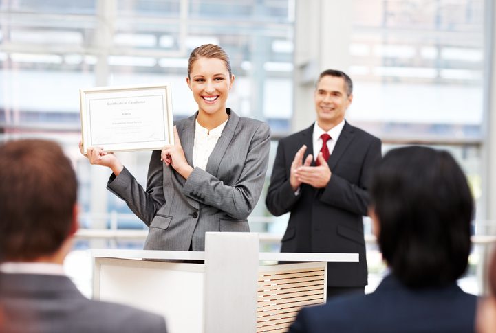 A person in a gray suit holds a framed certificate at a podium while another applauds in a bright room with seated attendees.