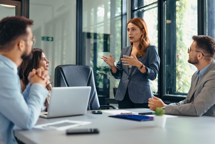 Professional woman leading a team discussion in a modern office setting, with three colleagues listening attentively around a conference table.