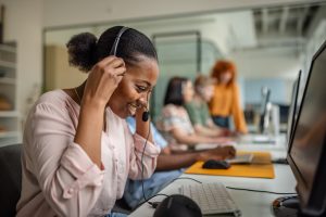Black woman wearing a headset and working at a computer in a modern office setting, with other team members collaborating in the background.