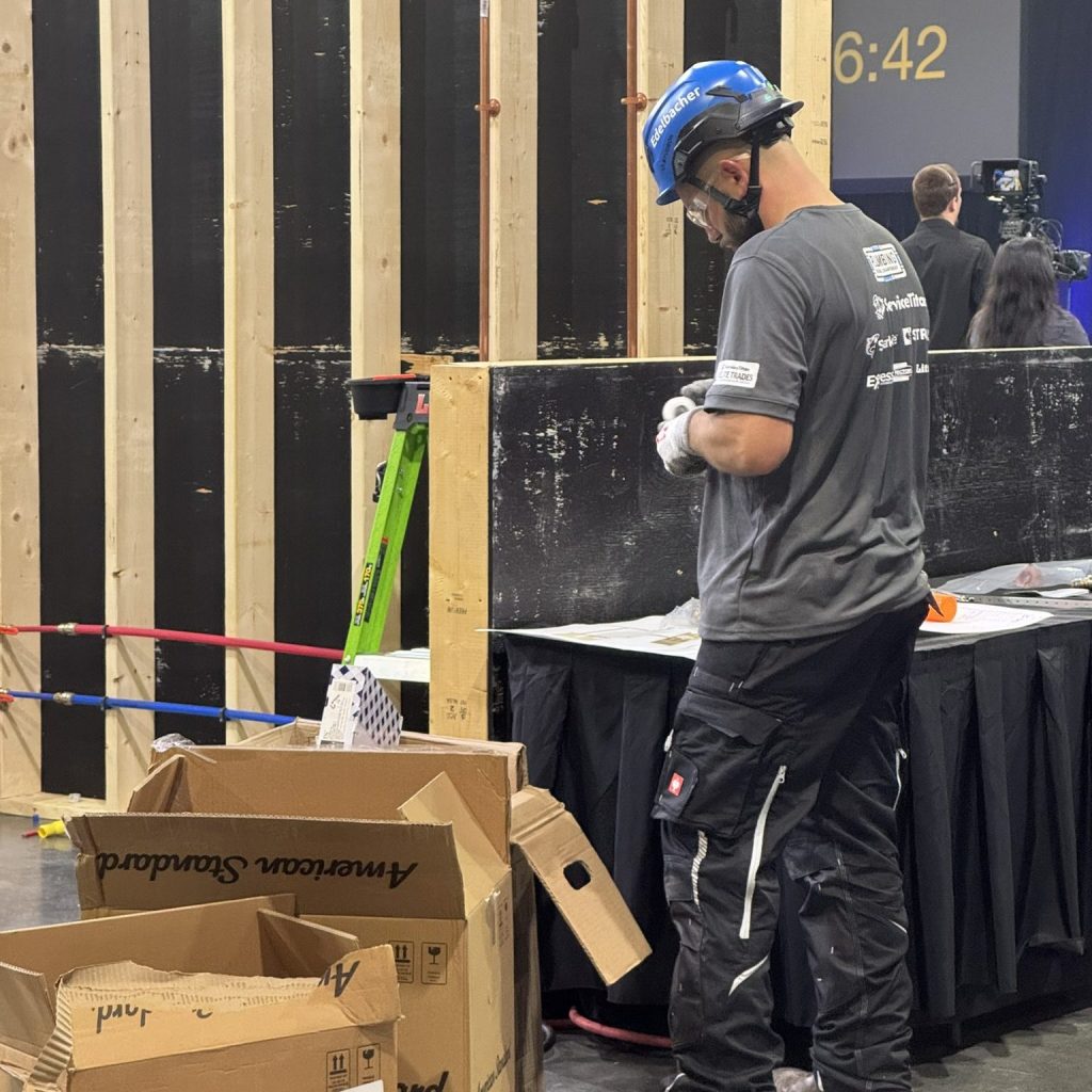 Competitor preparing plumbing components during the Elite Trades Championship Series, representing skilled trades excellence with tools and American Standard boxes.