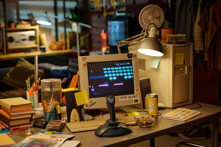 Photo of a cluttered desk with a retro computer setup. The CRT monitor displays a pixelated space-invader-style game with the text “Your score: 388” at the top. A sticker on the monitor reads “STAY ALERT!”. The desk has a joystick, keyboard, notebooks, pens in holders, sticky notes, a soda can, and a bowl of colorful candy. A desk lamp and small fan are attached to the computer tower. In the background, shelves with books, a radio, and a couch are visible, along with hanging clothes and a refrigerator.
