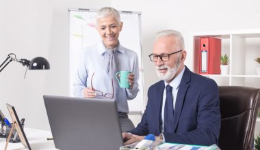Two older adults in a professional office setting smiling at a laptop screen. One stands holding a green coffee mug and red eyeglasses, while the other sits in a dark blue suit and tie. The desk features a laptop, lamp, framed photo, pen holder, and documents with charts. A flip chart, red binder, and potted plant are visible in the background.