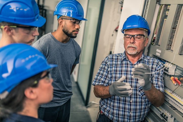 Photo of a group of people in an industrial training setting wearing blue safety helmets. One man in a checkered shirt and gloves is demonstrating electrical work near a control panel while others observe attentively.
