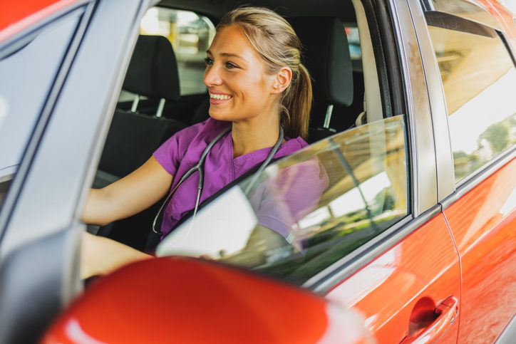 A nurse wearing scrubs and a stethoscope sits in the driver’s seat of a car, looking out the window as she prepares to travel for work.
