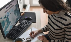 Person writing on paper at a desk with a computer screen showing “Job Search” and charts; keyboard, mouse, coffee cup, and eyeglasses nearby.