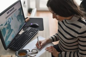 Person writing on paper at a desk with a computer screen showing “Job Search” and charts; keyboard, mouse, coffee cup, and eyeglasses nearby.