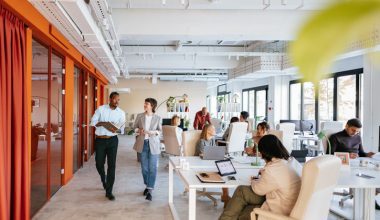 Photo of a modern open-plan office with several people working at desks and two individuals walking and talking in the foreground. The space features large windows, white walls, orange accents, and contemporary furniture.