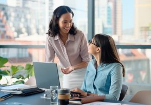 Two women collaborate at a modern office desk, with one standing and gesturing toward an open laptop while the other sits, listens, and learns. The workspace includes notebooks, a coffee cup, and a smartphone, and large windows behind them reveal a city skyline filled with natural light.