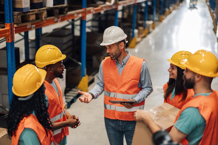 Photo of a group of workers in a warehouse wearing orange safety vests and yellow or white hard hats. A man in a white hard hat and orange vest stands at the center holding a tablet and speaking to the team. The others, including women and men, stand around him listening; one person on the right holds a cardboard box. Rows of shelves and inventory line the background.
