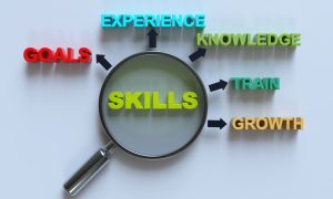 Photo of a magnifying glass highlighting the word “SKILLS” at the center, with surrounding words connected by arrows: “EXPERIENCE,” “KNOWLEDGE,” “TRAIN,” “GROWTH,” and “GOALS,” displayed on a light background in colorful, three‑dimensional text.