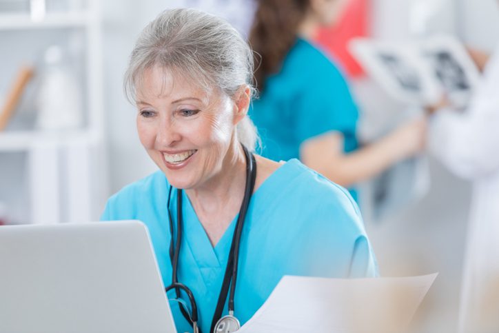 Cheerful healthcare leader reviewing digital records on a computer while completing paperwork at their desk.